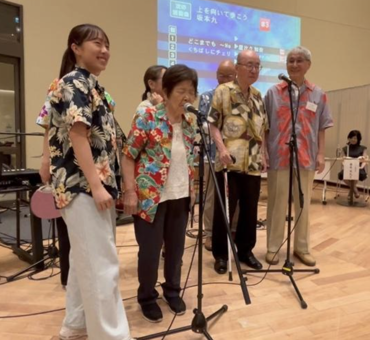 女性利用者がカラオケを歌っており、ヘルパーが寄り添っている写真