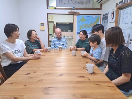 子供と大人の男女７名がコップを手に机を囲んでいるサロン風景写真。